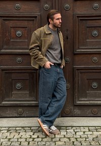 Veste en daim kaki portée sur un pull gris à côtes, associée à un jean bleu ample et des baskets marron à lacets blancs, debout devant des portes en bois.