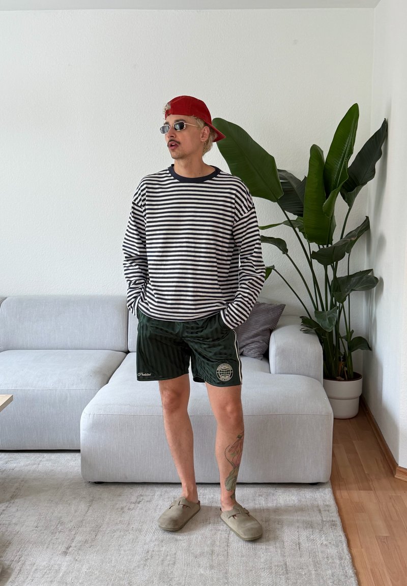 Personne portant une casquette rouge, une chemise à manches longues rayée, un short foncé et des chaussons se tient dans un salon près d'un canapé gris et d'une grande plante verte.