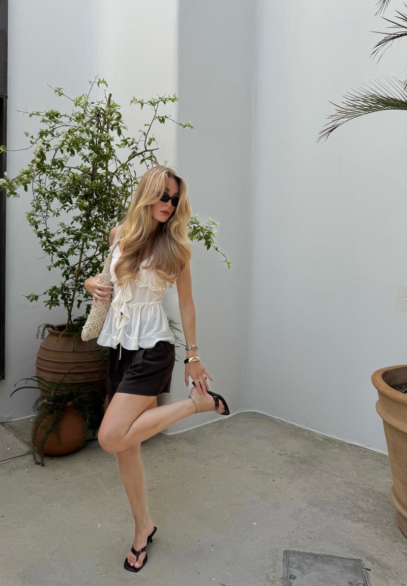 Top bianco a sbuffo e pantaloni corti pieghettati marrone scuro. Infradito nere. Capelli lunghi e mossi. Ambientazione naturale con pianta in vaso e muro.