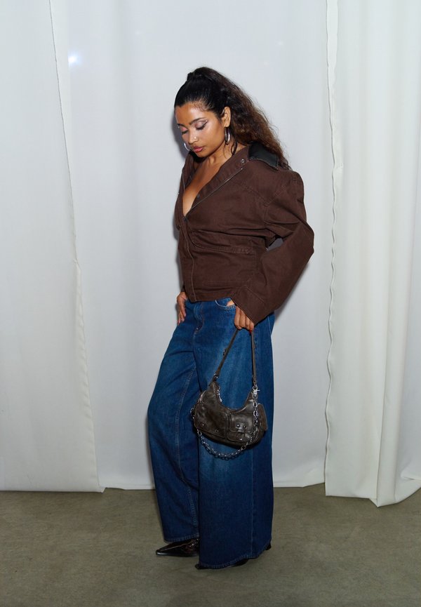 Mujer con el pelo largo, con una chaqueta marrón y vaqueros anchos azules, sostiene un pequeño bolso negro, de pie frente a una cortina blanca.