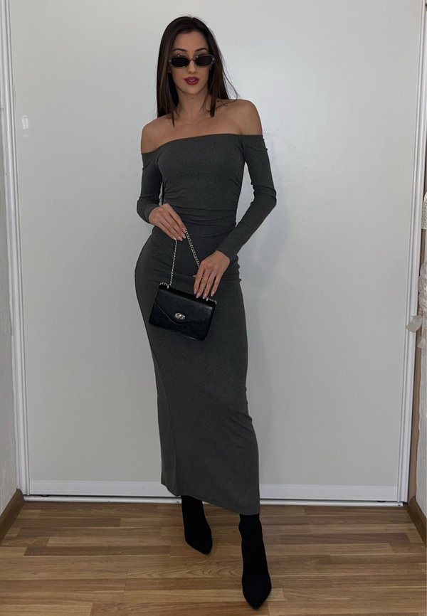 Vrouw draagt een nauwsluitende grijze jurk met blote schouders, zwarte enkellaarzen, een zonnebril en houdt een zwarte handtas met een kettingriem vast, staand op een houten vloer.