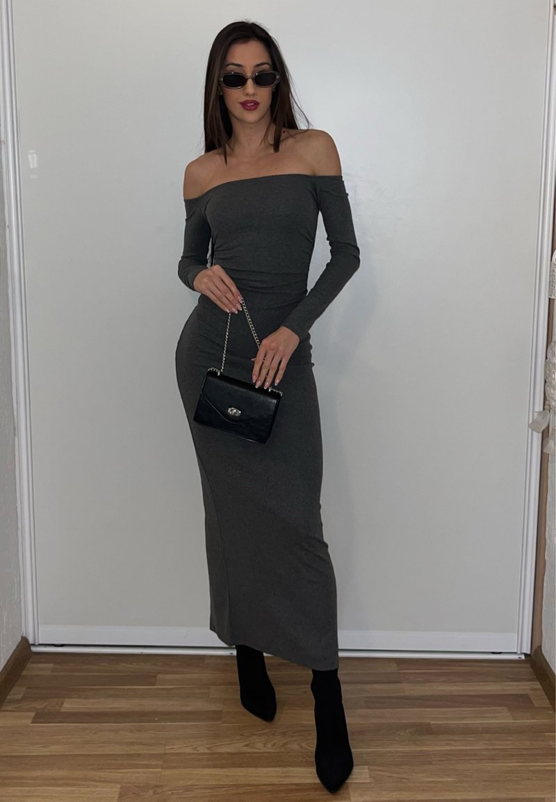 Femme portant une robe grise ajustée à épaules dénudées, des bottines noires, des lunettes de soleil, tenant un sac à main noir à bandoulière chaîne, debout sur un sol en bois.