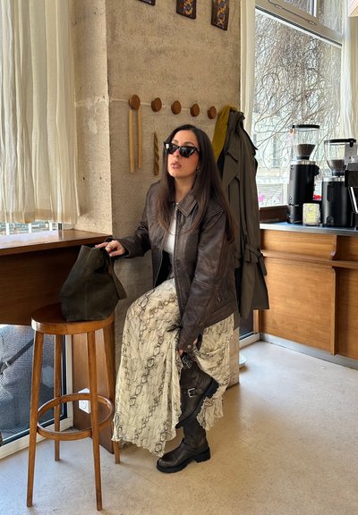 Chaqueta de cuero marrón oscuro, falda larga crema con estampado, gafas de sol negras y botas negras. Bolsa de ante marrón al lado de un taburete de madera.