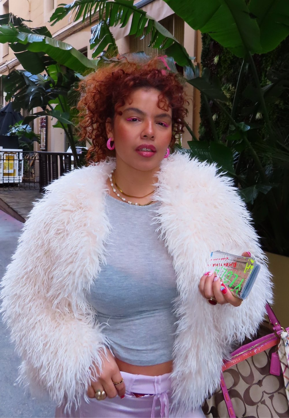 Jeune femme aux cheveux roux bouclés portant une veste blanche duveteuse, des boucles d'oreilles roses, tenant une coque de téléphone colorée près de grandes plantes vertes en plein air.