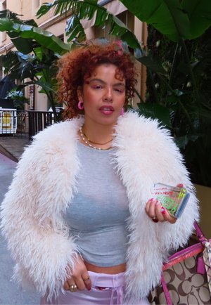 Joven con cabello rizado y rojo, vistiendo una chaqueta blanca y esponjosa, pendientes rosas y sosteniendo una funda de móvil colorida cerca de grandes plantas verdes al aire libre.