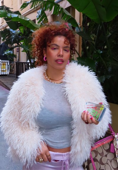 Joven con cabello rizado y rojo, vistiendo una chaqueta blanca y esponjosa, pendientes rosas y sosteniendo una funda de móvil colorida cerca de grandes plantas verdes al aire libre.