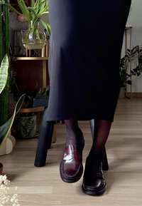 Personne portant des bas noirs, une longue jupe noire et des mocassins en cuir brun foncé, assise à l'intérieur près de plantes et d'un sol en bois.