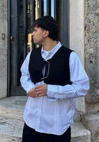 Jeune homme aux cheveux foncés portant une chemise rayée et un gilet noir, debout près d'un mur en pierre et d'une porte vitrée, regardant vers la gauche.