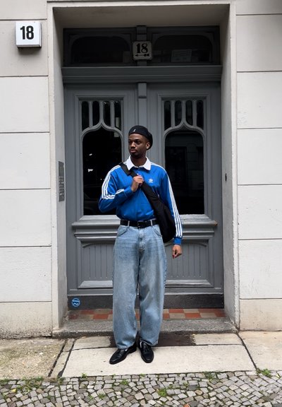Un joven se encuentra frente a unas puertas dobles grises con el número 18, vistiendo un sudadera azul de Adidas, jeans holgados, zapatos negros y una boina negra.