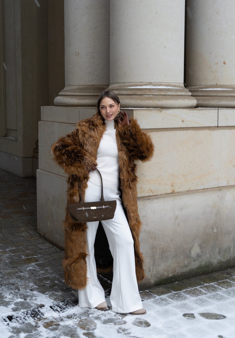 Manteau en fausse fourrure marron, pull ajusté blanc et pantalon blanc à jambes larges. Tenant un sac à main marron avec des détails métalliques, debout près de colonnes beiges.