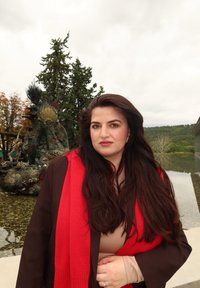 Kvinna i brun kappa och röd scarf står nära en reflekterande vattenspegel, med träd och en dekorativ skulptur i bakgrunden.