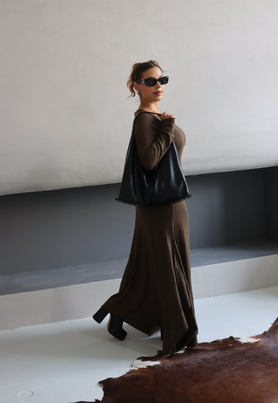 Mujer con un vestido largo marrón y botas de tacón negras lleva un gran bolso negro, usa gafas de sol y está de pie en el interior cerca de una alfombra marrón.