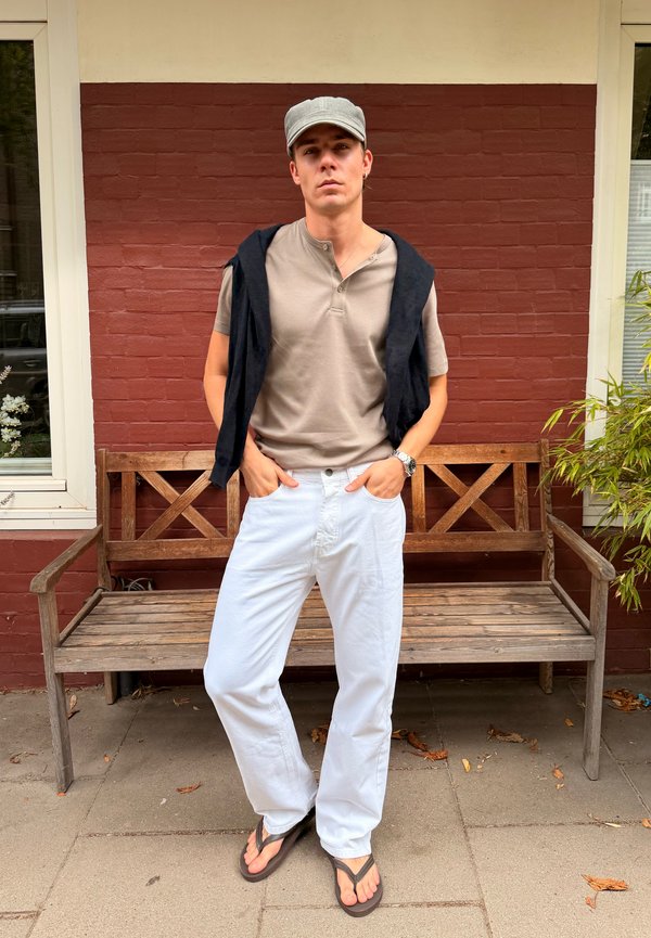 Jeune homme portant une chemise beige, un pantalon blanc, une casquette grise, un pull noir sur les épaules et des tongs, debout devant un banc en bois et un mur en briques rouges.