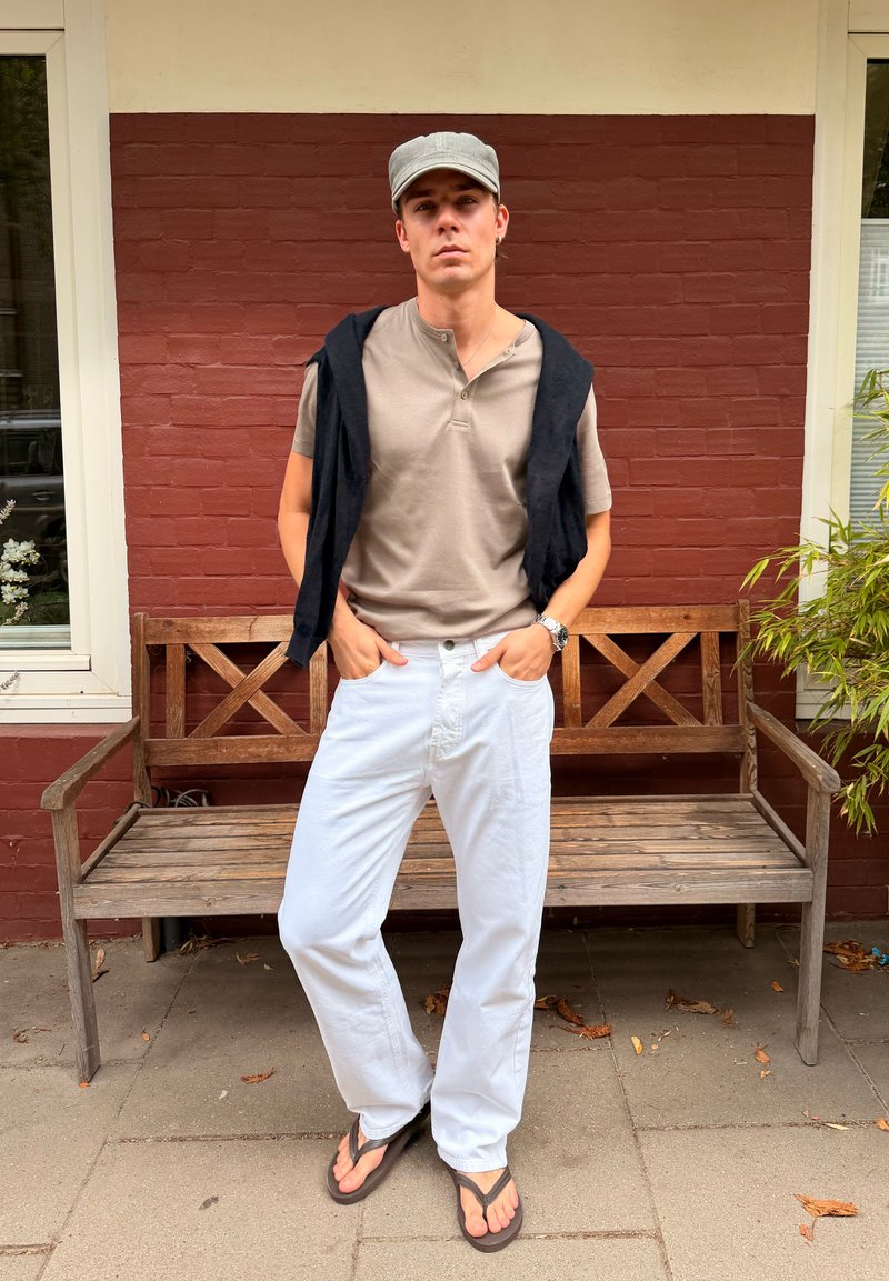 Jeune homme portant une chemise beige, un pantalon blanc, une casquette grise, un pull noir sur les épaules et des tongs, debout devant un banc en bois et un mur en briques rouges.