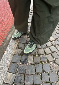 Zielone buty sportowe z teksturowaną siateczką na cholewce i gumową podeszwą, w zestawieniu z luźnymi ciemnymi spodniami, umieszczone na bruku.