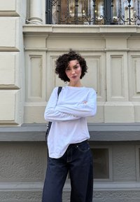 Vrouw met kort krullend haar, gekleed in een witte blouse met lange mouwen en donkere jeans, staat met gekruiste armen voor een beige versierde muur.