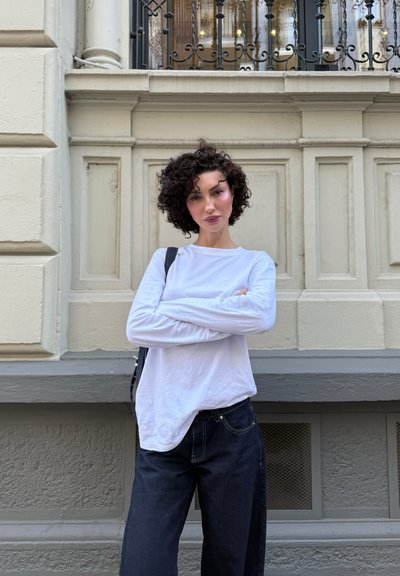 Mujer con cabello rizado corto, vestida con una camisa blanca de manga larga y jeans oscuros, se encuentra de brazos cruzados frente a una pared beige ornamentada.