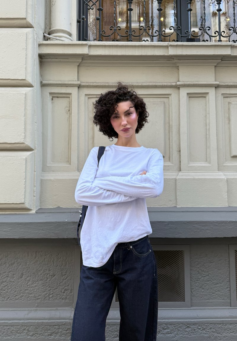 Donna con capelli corti e ricci, che indossa una camicia bianca a maniche lunghe e jeans scuri, si trova con le braccia incrociate di fronte a una parete beige ornata.