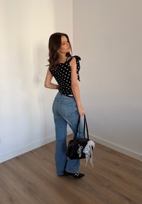 Femme portant un haut noir à pois et un jean bleu, tenant un sac à main noir avec un foulard en dentelle blanche, debout sur un sol en bois près d'un mur blanc.