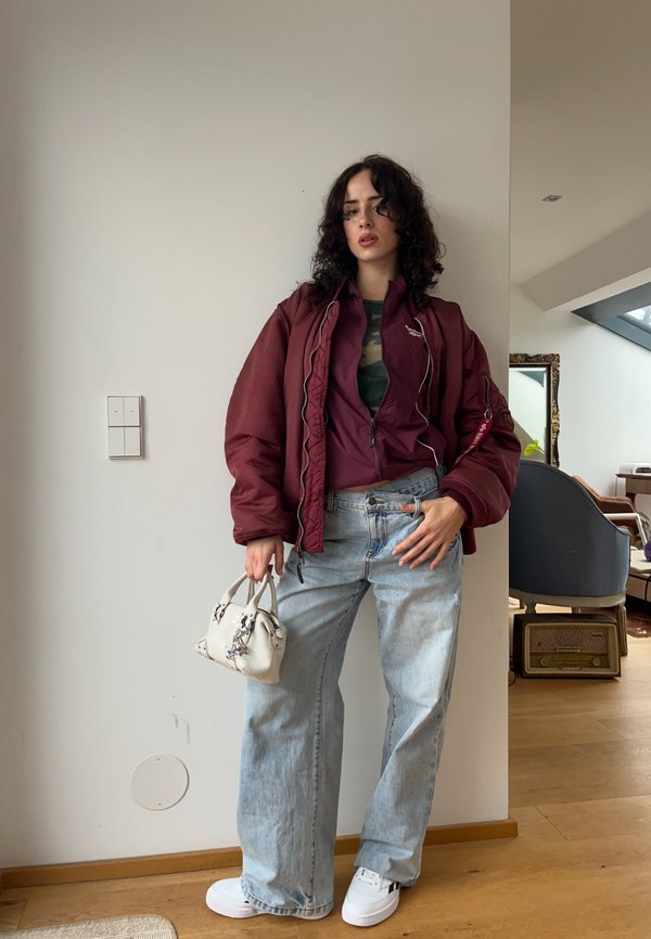 Woman with curly dark hair wearing an oversized maroon jacket, camouflage shirt, loose light blue jeans, white trainers, holding a small white handbag indoors.