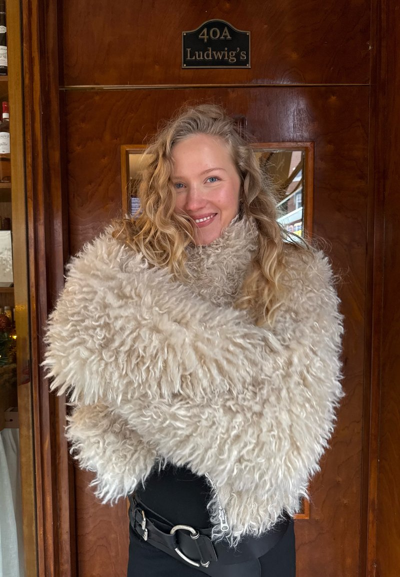 Fluffiger, cremefarbener Fellschal aus langen, strukturierten Fasern; um die Schultern drapiert; kombiniert mit einem schwarzen Gürtel und dunkler Kleidung.