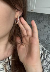 Femme ajustant une boucle d'oreille créole en or ornée de pierres précieuses transparentes, main près de l'oreille, portant une chemise à carreaux, avec un dosseret en marbre et une armoire blanche en arrière-plan.