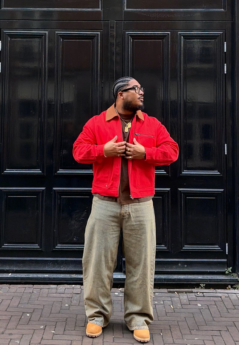 Homme aux cheveux tressés et portant des lunettes ajuste sa veste rouge, debout devant de grandes portes noires à panneaux sur une rue pavée.