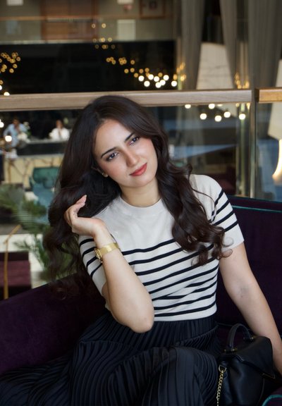 Joven mujer con cabello oscuro y largo, vestida con una camisa de rayas y una falda negra, sentada en un sofá morado, descansando su mano en su cabello en el interior.