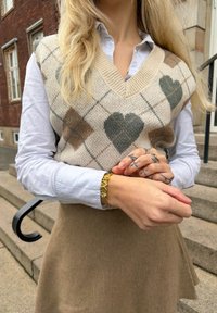 Veste beige en tricot avec motifs de cœurs, chemise boutonnée bleu clair et jupe beige. Comprend un bracelet en or avec des accents en forme de cœur.
