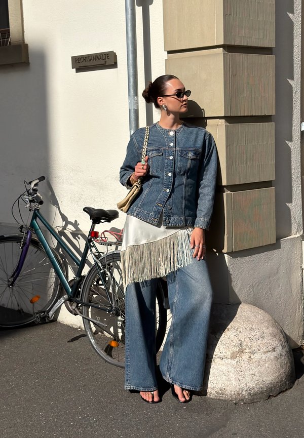Mujer con chaqueta vaquera y vaqueros anchos, descalza, apoyada contra la esquina de un edificio junto a una bicicleta estacionada en una calle soleada.