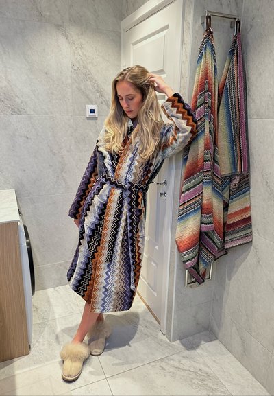 Mujer en un albornoz con estampado y zapatillas de felpa apoyada contra una puerta blanca en un baño de baldosas de mármol, con toallas del mismo estampado colgando cerca.