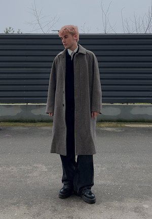 Joven con cabello rosa lleva un abrigo largo de cuadros sobre un conjunto negro, de pie en un área pavimentada con una cerca gris oscura al fondo.