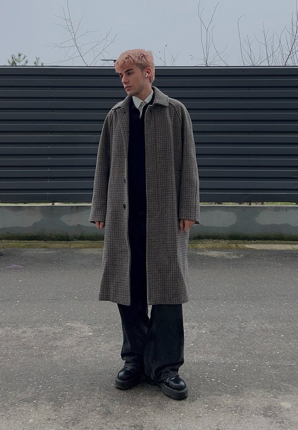 Ung person med rosa hår har på seg en lang rutete frakk over et svart antrekk, stående på et belegningsområde med et mørkegrått gjerde i bakgrunnen.