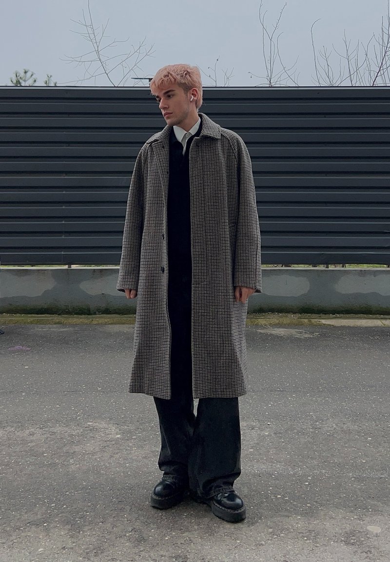 Jeune personne aux cheveux roses portant un long manteau à carreaux par-dessus une tenue noire, debout sur une surface pavée avec une clôture gris foncé en arrière-plan.