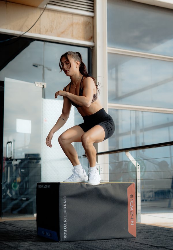Kvinde i sportstøj udfører hop-squats på en blød plyoboks inde i et fitnesscenter med store vinduer i baggrunden.