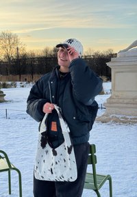 Personne souriante en plein air au coucher du soleil dans un parc enneigé, portant une veste bleu marine, une casquette de baseball rayée et tenant un sac blanc à taches noires.