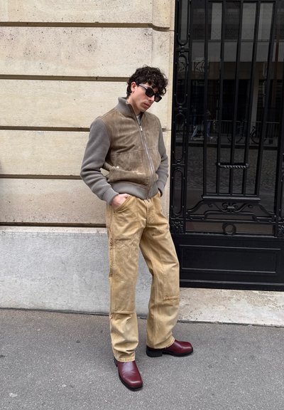 Hombre con gafas de sol, chaqueta y pantalones beige, y zapatos rojos apoyado contra una pared de piedra cerca de una puerta de metal negro.