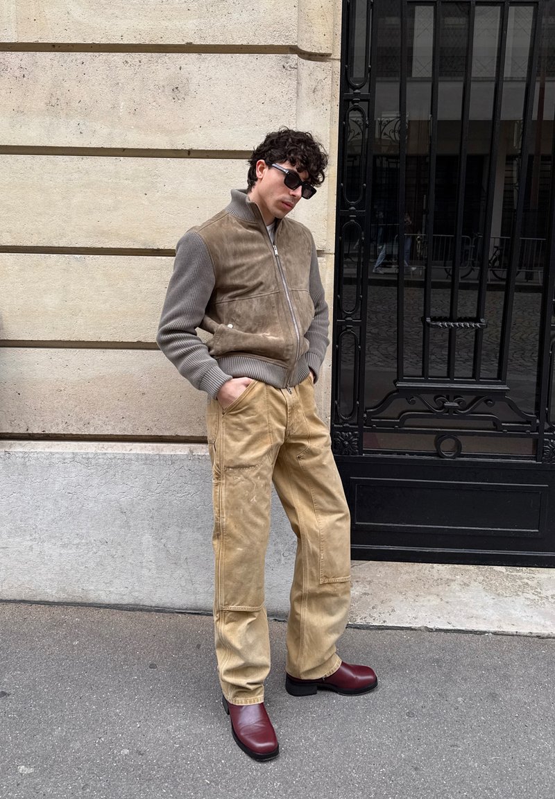 Man wearing sunglasses, beige jacket and pants, and red shoes leaning against a stone wall near a black metal gate.