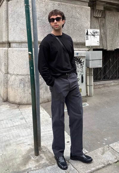 Hombre con gafas de sol, camisa de manga larga negra, pantalones gris oscuro y zapatos negros, de pie en una acera de la ciudad con cajas de servicios cubiertas de graffiti.