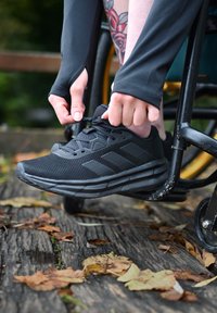 Černé sportovní boty s texturovaným síťovým svrškem a polstrovanou podrážkou, které se svazují rukama. Rám invalidního vozíku částečně viditelný v pozadí.