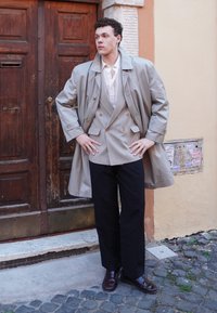 Beige trenchcoat over en grå dobbeltbrystet jakkesæt, parret med sorte bukser og brune sko. Baggrunden indeholder trædøre og en væg.