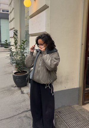 Una persona con una chaqueta bomber gris y pantalones anchos negros ajusta sus gafas de sol mientras lleva puestos unos auriculares, de pie junto a una planta en maceta.