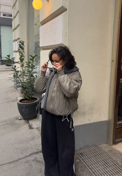 Una persona con una chaqueta bomber gris y pantalones anchos negros ajusta sus gafas de sol mientras lleva puestos unos auriculares, de pie junto a una planta en maceta.