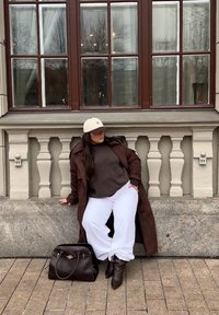 Bruine trenchcoat over een donkere trui, witte wijde broekspijpen en bruine leren laarzen. Een zwarte handtas ligt ernaast. Raam met bruine kozijnen op de achtergrond.