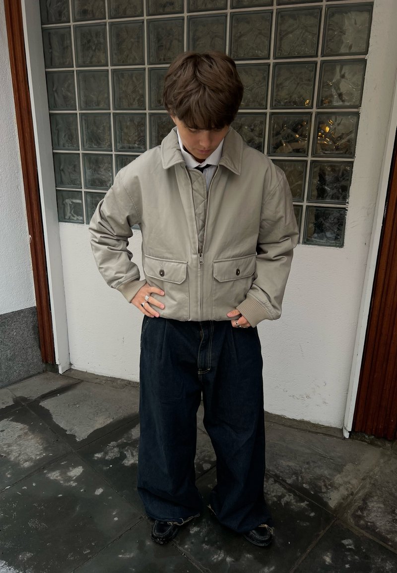Jeune personne portant une veste beige et un pantalon large foncé, debout à l'intérieur sur un sol carrelé foncé près d'une fenêtre en verre.