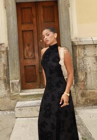 Robe à motifs floraux noirs avec un design de cou halter. Tissu lisse et élégant tombe jusqu'au sol. Des accessoires dorés complètent le look.