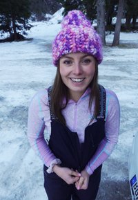 Strikket rosa og lilla lue, mønstret langermet topp i lys rosa, svarte kjeledresser med justerbare stropper, og snødekt bakgrunn.