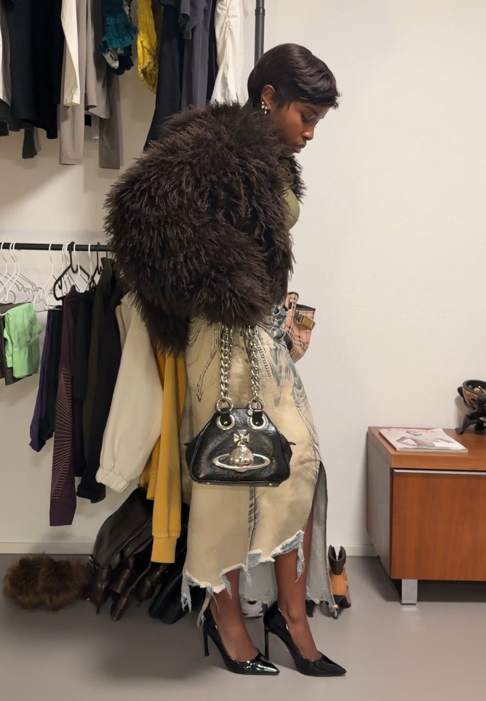 Modèle portant un manteau en fourrure shaggy marron foncé, un haut olive, une jupe en denim déchirée et des talons en patin noir, tenant un petit sac à main noir brillant.