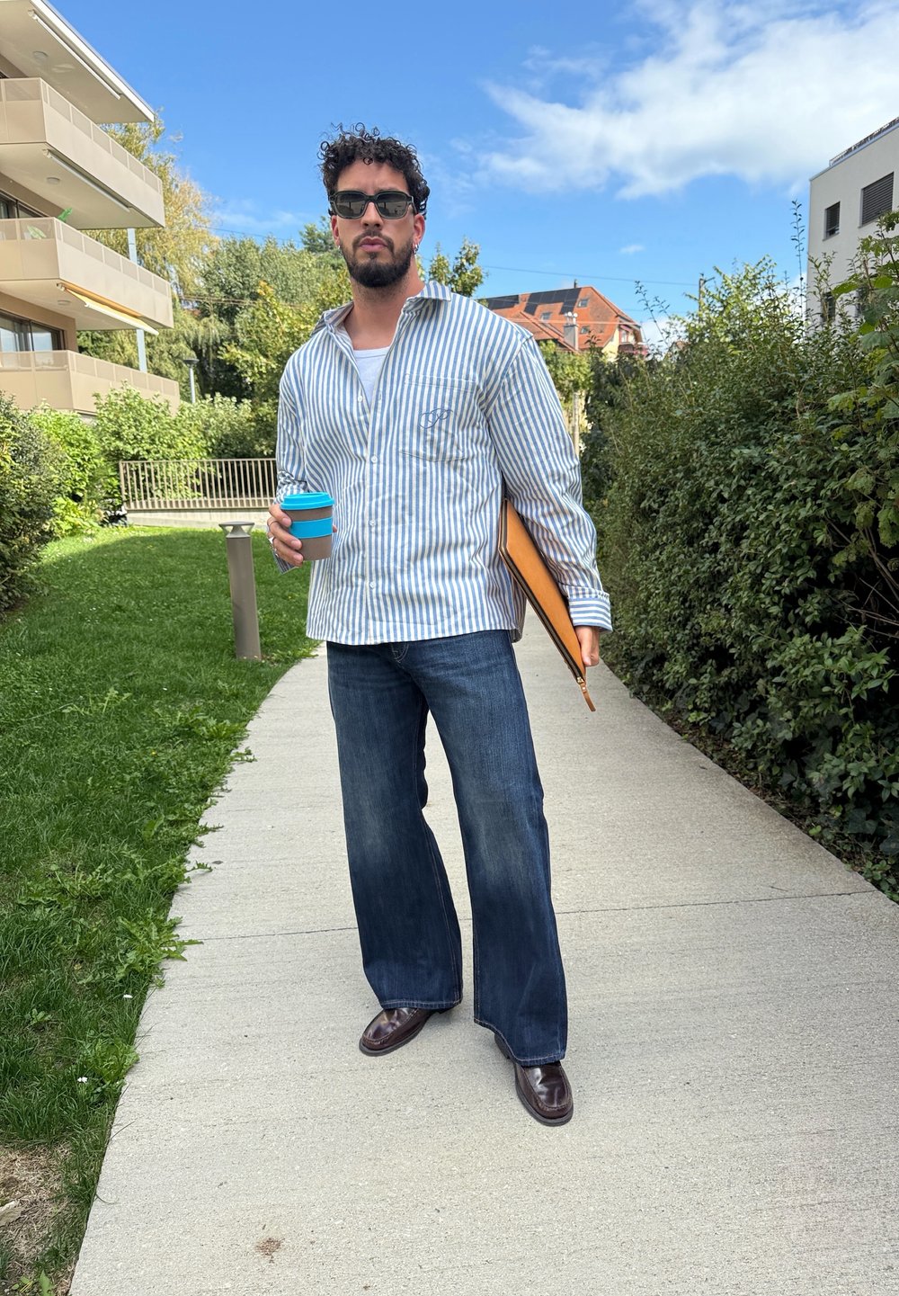 Camisa de botões às riscas em azul e branco, jeans escuros, sapatos castanhos, segurando um copo de café castanho com uma tampa azul e um bloco de notas.