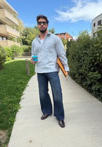 Camisa de botões às riscas em azul e branco, jeans escuros, sapatos castanhos, segurando um copo de café castanho com uma tampa azul e um bloco de notas.